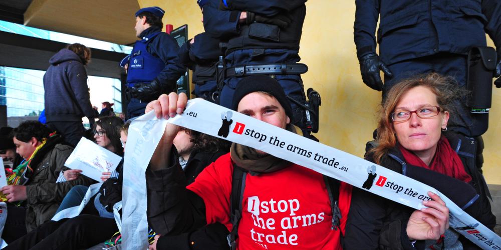 actiefoto met een aantal activisten aan een gebouw waarvoor de politie staat. Een activist houdt een lint met I Stop The Arms Trade vast. 