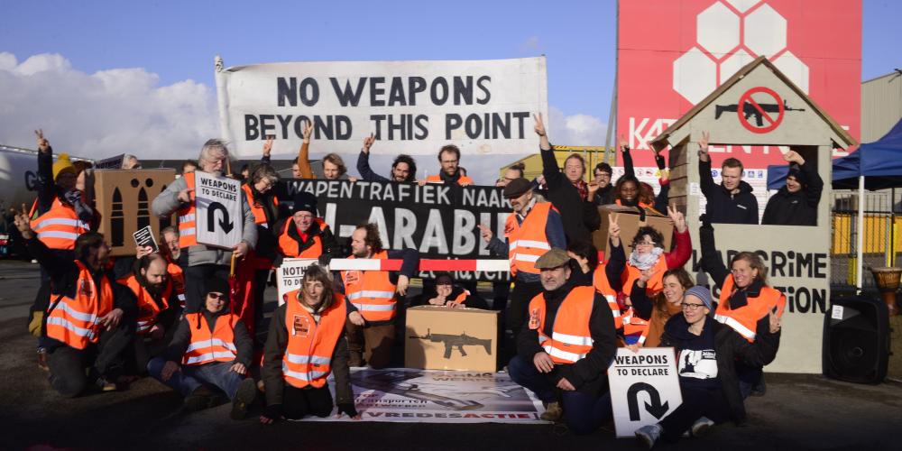 foto van protest met activisten, douanehuisje met bareel en banners: No Weapons Beyond this Point + Stop de wapentrafiek naar Saoedi-Arabië