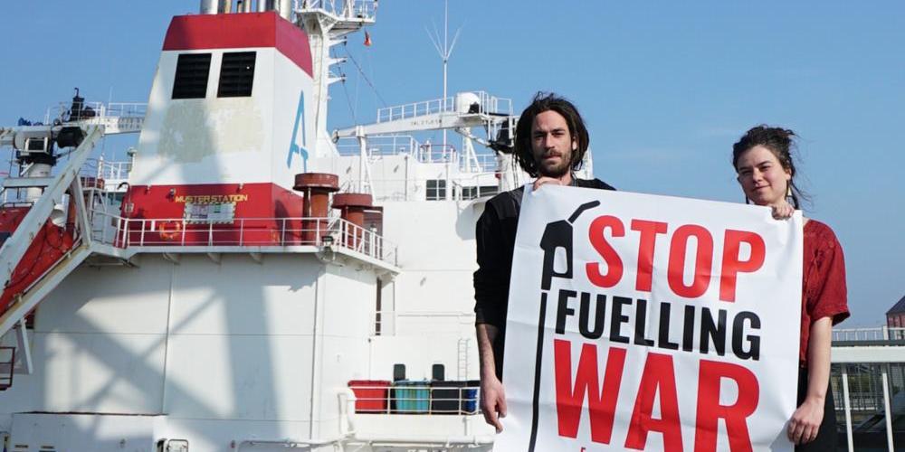 Twee mensen staan aan een sluis bij een Russische olietanker. Ze houden een banner vast waarop staat "Stop Fuelling War".