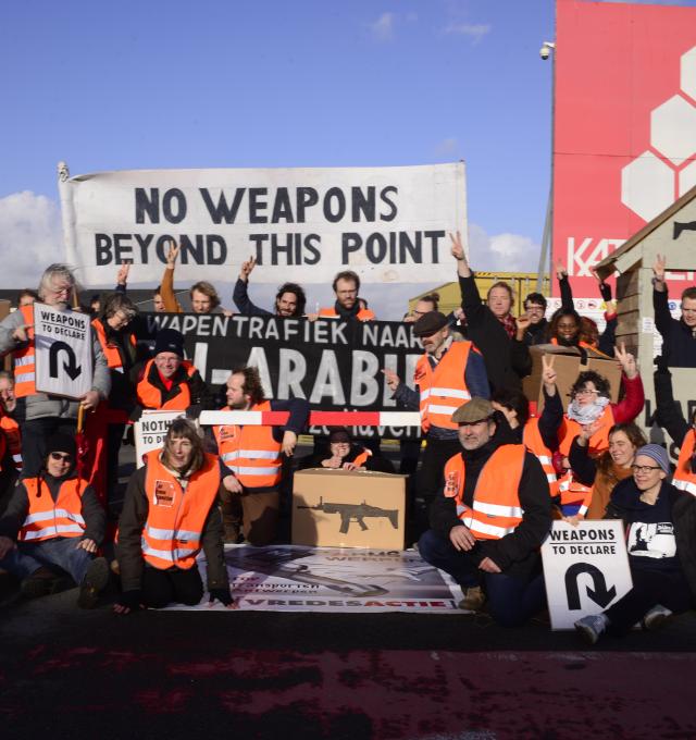 foto van protest met activisten, douanehuisje met bareel en banners: No Weapons Beyond this Point + Stop de wapentrafiek naar Saoedi-Arabië
