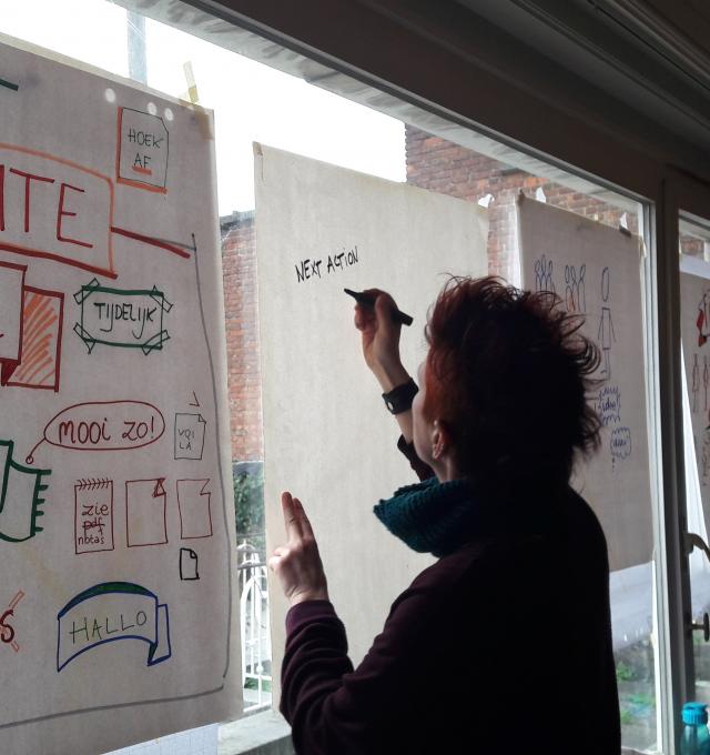 2 mensen schrijven op flipcharts op een raam. 1 flipchart is al volledig beschreven met kadertjes, tekstjes en andere dingen.